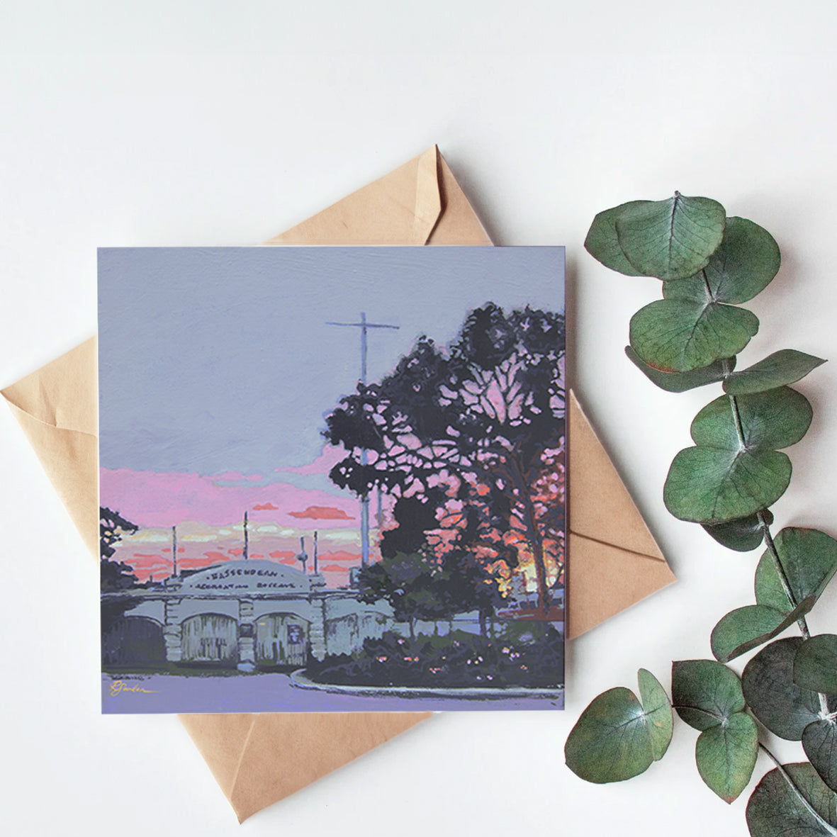 Greeting card with a scenic illustration of a building and trees, placed on a white surface with a brown envelope and green leaves.